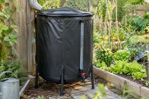 Récupérateur d’eau de pluie pliable noir avec couvercle grillagé et tube gradué près d’un potager.